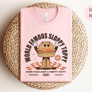 May include: A pink t-shirt featuring a cartoon burger wearing a peace sign and the text "World Famous Sloppy Toppy" and "More Than Just A Pretty Patty".