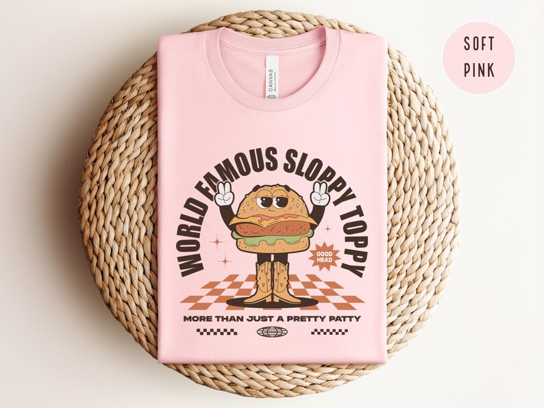 May include: A pink t-shirt featuring a cartoon burger wearing a peace sign and the text "World Famous Sloppy Toppy" and "More Than Just A Pretty Patty".