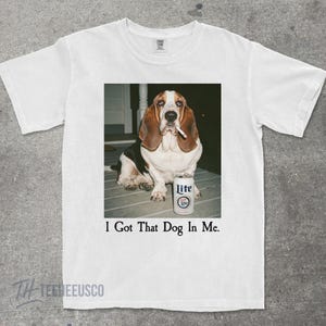 May include: White t-shirt featuring a Basset Hound with a cigarette in its mouth, next to a can of Lite beer. The text "I Got That Dog In Me" is printed below the image. The shirt has a crew neck and short sleeves.