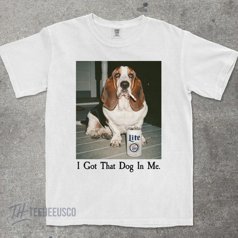 May include: White t-shirt featuring a Basset Hound with a cigarette in its mouth, next to a can of Lite beer. The text "I Got That Dog In Me" is printed below the image. The shirt has a crew neck and short sleeves.
