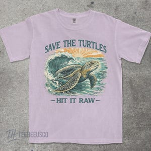 May include: A light purple t-shirt with a graphic of a sea turtle swimming in the ocean. The text "SAVE THE TURTLES" is above the turtle, and "- HIT IT RAW -" is below. The shirt has short sleeves and a crew neck.
