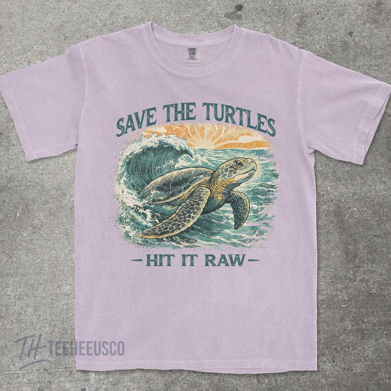 May include: A light purple t-shirt with a graphic of a sea turtle swimming in the ocean. The text "SAVE THE TURTLES" is above the turtle, and "- HIT IT RAW -" is below. The shirt has short sleeves and a crew neck.
