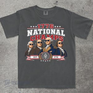 May include: Dark gray t-shirt featuring a graphic with the text "1776 NATIONAL CHAMPS" and an illustration of four historical figures wearing sunglasses. The design includes stars, a banner, and the year 1776.