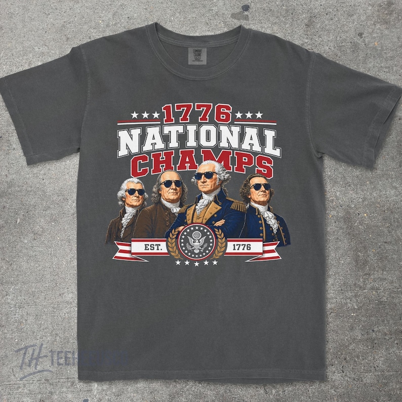 May include: Dark gray t-shirt featuring a graphic with the text "1776 NATIONAL CHAMPS" and an illustration of four historical figures wearing sunglasses. The design includes stars, a banner, and the year 1776.