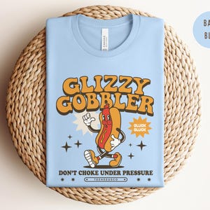 K&ouml;nnte beinhalten: Hellblaues T-Shirt mit einem Retro-Cartoon-Hotdog-Motiv. Der Text "Glizzy Gobbler" ist in Orange gehalten, darunter der Satz "Don't Choke Under Pressure". Das T-Shirt liegt gefaltet auf einer gewebten Matte.