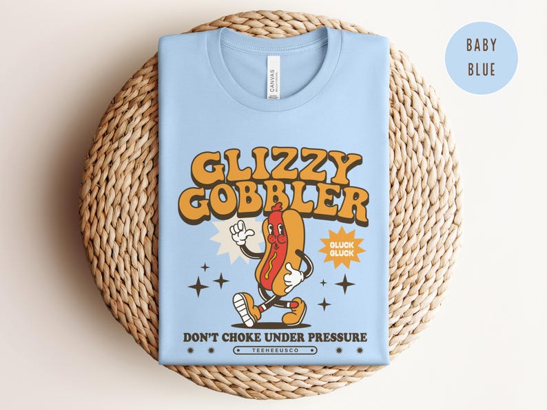 K&ouml;nnte beinhalten: Hellblaues T-Shirt mit einem Retro-Cartoon-Hotdog-Motiv. Der Text "Glizzy Gobbler" ist in Orange gehalten, darunter der Satz "Don't Choke Under Pressure". Das T-Shirt liegt gefaltet auf einer gewebten Matte.