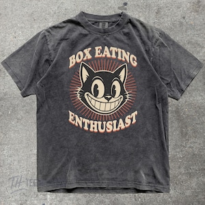 May include: A charcoal gray short-sleeve t-shirt featuring a graphic of a smiling cat face centered within a sunburst design. The text "BOX EATING ENTHUSIAST" is printed in a curved, cream-colored font above and below the cat illustration.