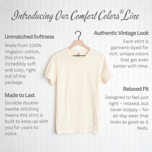 May include: Cream-colored Comfort Colors t-shirt hanging on a wooden hanger. The shirt is made of ringspun cotton and has a relaxed fit. Text on the image reads "Introducing Our Comfort Colors Line."