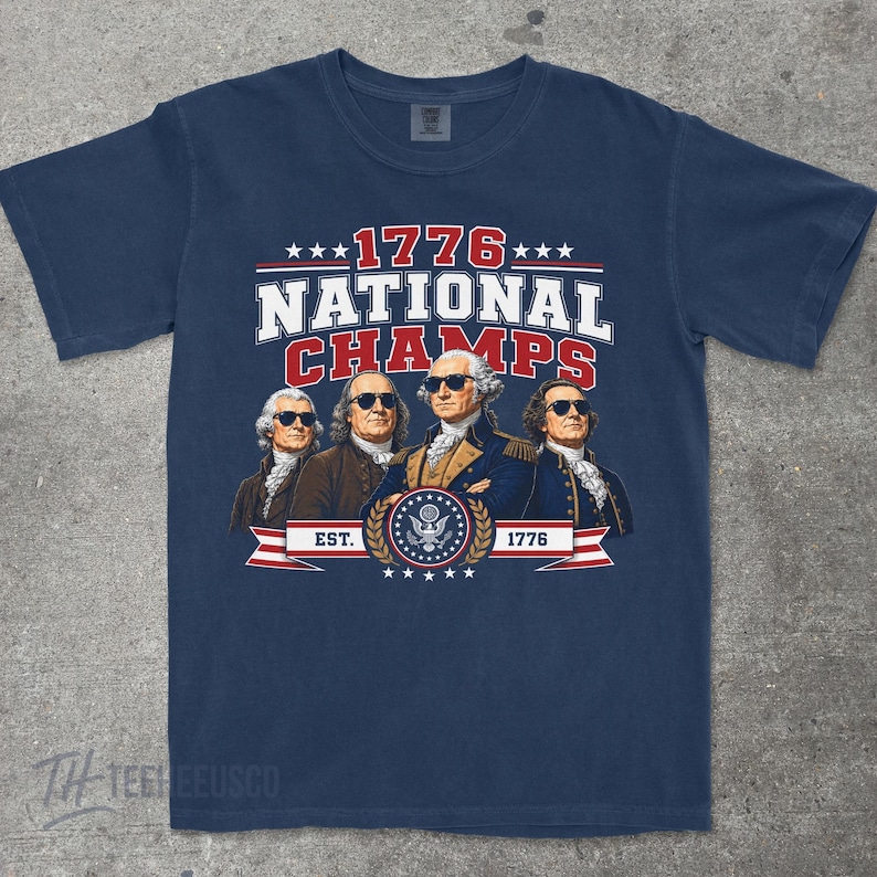May include: Navy blue t-shirt with a graphic featuring four historical figures wearing sunglasses. The text reads "1776 NATIONAL CHAMPS" with "EST. 1776" below a patriotic emblem. The shirt is a classic crew neck.