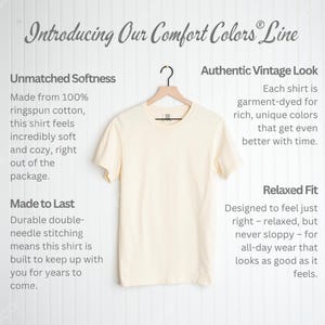 May include: Cream-colored Comfort Colors t-shirt hanging on a wooden hanger. The shirt is displayed against a white background with text that reads "Introducing Our Comfort Colors Line". The shirt is made of ringspun cotton.