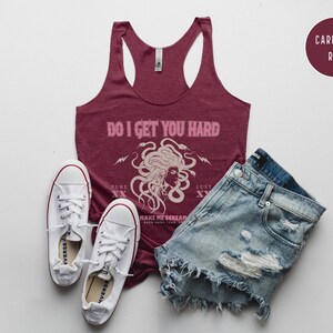 May include: A burgundy tank top with the text "DO I GET YOU HARD" and a Medusa graphic, alongside distressed denim shorts and white trainers. A "CARDINAL RED" colour swatch is also visible in the image.