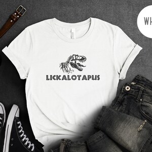 Może przedstawiać: Biała koszulka z czarnym nadrukiem szkieletu dinozaura i tekstem "LICKALOTAPUS" na przodzie.