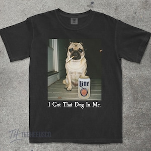Pu&ograve; includere: T-shirt nero con un'immagine umoristica di un carlino con una sigaretta in bocca e una lattina di birra Lite. Il testo "I Got That Dog In Me" &egrave; stampato sotto. La maglietta &egrave; di colore scuro.