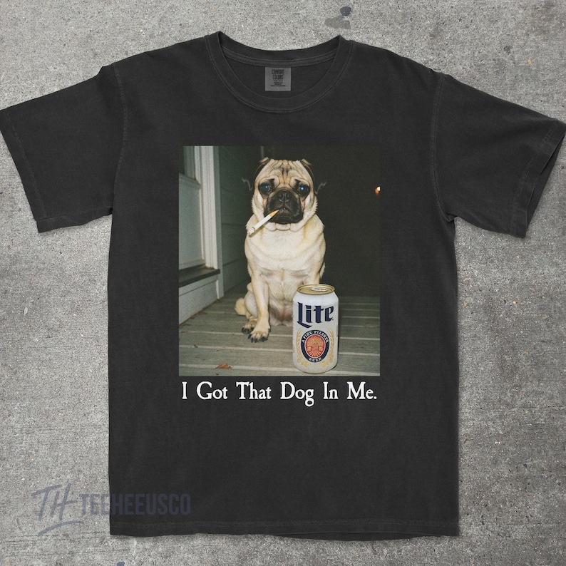 Pu&ograve; includere: T-shirt nero con un'immagine umoristica di un carlino con una sigaretta in bocca e una lattina di birra Lite. Il testo "I Got That Dog In Me" &egrave; stampato sotto. La maglietta &egrave; di colore scuro.