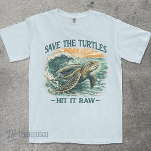 May include: Light blue t-shirt with a graphic of a sea turtle swimming in the ocean. The text "SAVE THE TURTLES" is above the turtle, and "- HIT IT RAW -" is below. The design includes a sunset and waves.