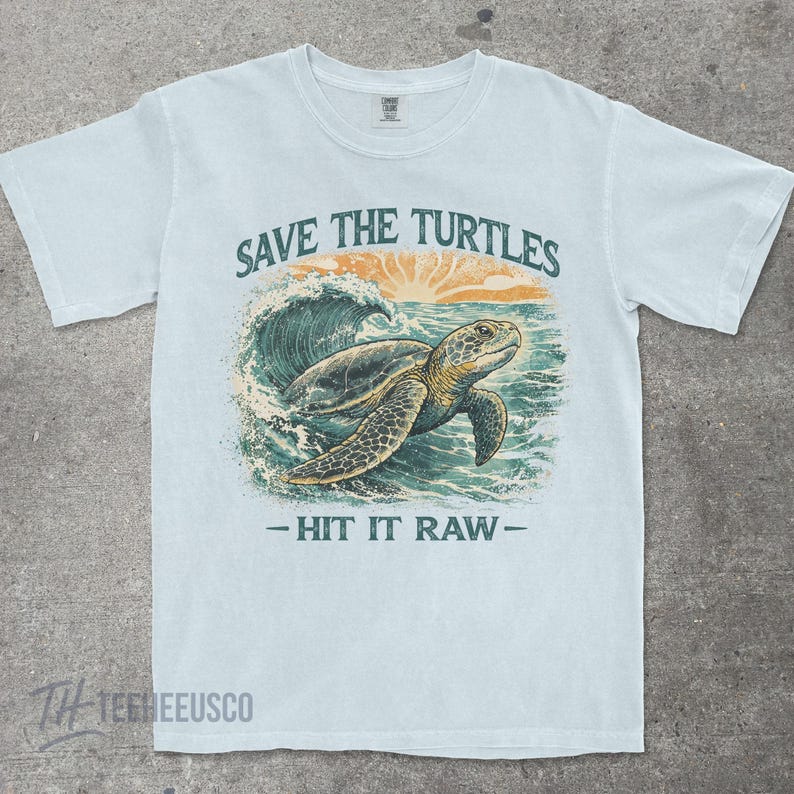 May include: Light blue t-shirt with a graphic of a sea turtle swimming in the ocean. The text "SAVE THE TURTLES" is above the turtle, and "- HIT IT RAW -" is below. The design includes a sunset and waves.