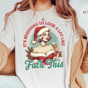 Pu&ograve; includere: T-shirt color crema con un'illustrazione in stile vintage di una donna con un cappello da Babbo Natale che tiene in mano un bicchiere di vino. Il testo recita "It's beginning to look a lot like Fuck This" in rosso e verde.