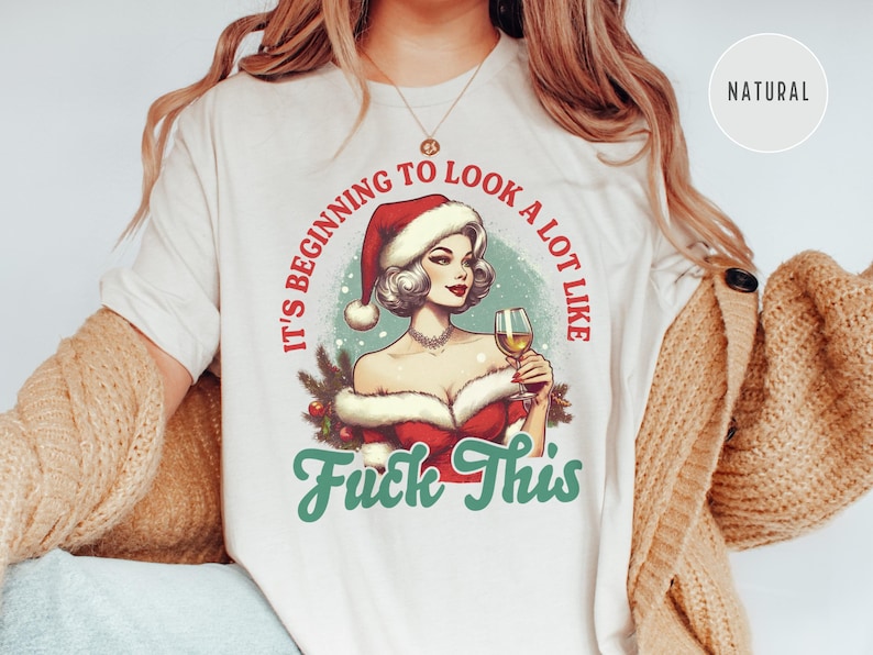 Pu&ograve; includere: T-shirt color crema con un'illustrazione in stile vintage di una donna con un cappello da Babbo Natale che tiene in mano un bicchiere di vino. Il testo recita "It's beginning to look a lot like Fuck This" in rosso e verde.