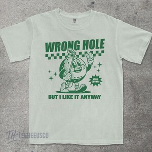 May include: Light green t-shirt with a green cartoon golf ball character holding a golf club. The text "WRONG HOLE" is above the character, and "BUT I LIKE IT ANYWAY" is below. The shirt has a vintage, distressed look.