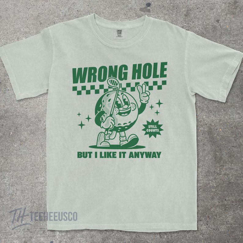 May include: Light green t-shirt with a green cartoon golf ball character holding a golf club. The text "WRONG HOLE" is above the character, and "BUT I LIKE IT ANYWAY" is below. The shirt has a vintage, distressed look.
