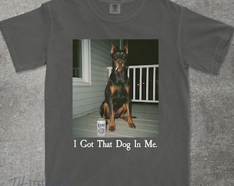 Puede incluir: Camiseta gris oscuro con una foto de un Doberman Pinscher con un cigarrillo en la boca, junto a una lata de cerveza Lite. El texto "I Got That Dog In Me" está impreso debajo.