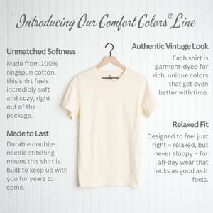 May include: A cream-colored t-shirt hangs on a wooden hanger. The text "Introducing Our Comfort Colors Line" is at the top. The shirt is described as having unmatched softness, an authentic vintage look, and a relaxed fit.