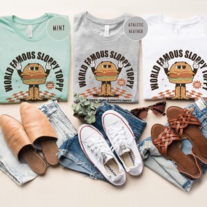 May include: Three t-shirts featuring a vintage cartoon burger design. The shirts are in mint, heather grey, and white. The text on the shirts reads "World Famous Sloppy Toppy".