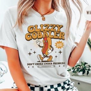 K&ouml;nnte beinhalten: Wei&szlig;es T-Shirt mit Retro-Grafik. Das Design zeigt eine Cartoon-Hotdog-Figur mit dem Text "GLIZZY GOBBLER" in Orange und Braun sowie dem Satz "DON'T CHOKE UNDER PRESSURE".