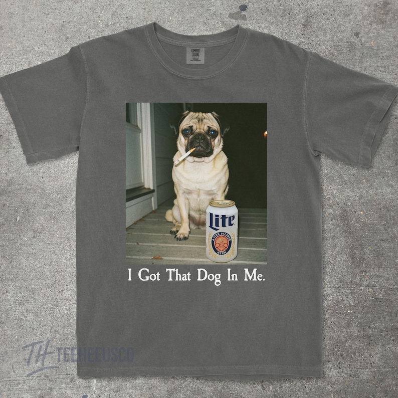 Pu&ograve; includere: T-shirt grigio scuro con una foto di un carlino con una sigaretta e una lattina di birra Lite. Il testo "I Got That Dog In Me" &egrave; stampato sotto. Un capo d'abbigliamento casual.