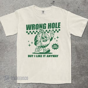 May include: Off-white t-shirt with a green graphic of a golf ball character holding a golf club and giving a peace sign. Text above reads "WRONG HOLE" and below "BUT I LIKE IT ANYWAY". The shirt has a crew neck and short sleeves.