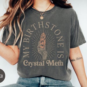 May include: A dark gray t-shirt with the text "My Birthstone Is Crystal Meth" in a vintage style font. The text is surrounded by a circle with a stylized crystal inside.