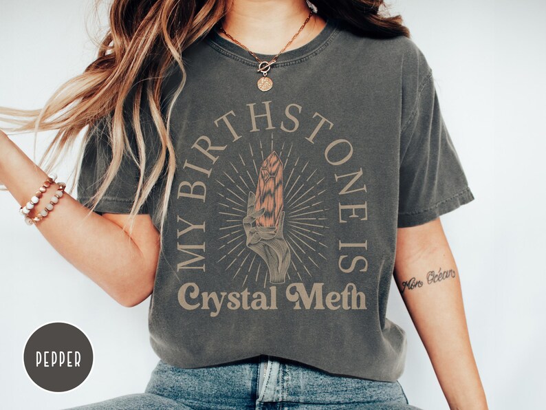 May include: A dark gray t-shirt with the text "My Birthstone Is Crystal Meth" in a vintage style font. The text is surrounded by a circle with a stylized crystal inside.