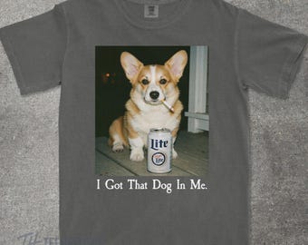 May include: Dark gray t-shirt featuring a graphic of a corgi dog with a cigarette in its mouth and a can of Lite beer. The text "I Got That Dog In Me" is printed below the image. The shirt is made of a soft material.
