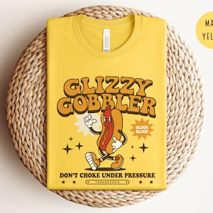 K&ouml;nnte beinhalten: Maisgelbes T-Shirt mit einem Retro-Cartoon-Hotdog-Motiv. Der Text lautet "Glizzy Gobbler" und "Don't Choke Under Pressure". Das Design umfasst eine laufende Hotdog-Figur und Sternakzente.