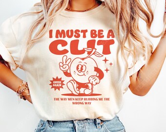 May include: Cream-colored sweatshirt with a red graphic. The graphic features an anthropomorphic orange with the text "I MUST BE A CLIT" above it. Below the orange is the text "THE WAY MEN KEEP RUBBING ME THE WRONG WAY."