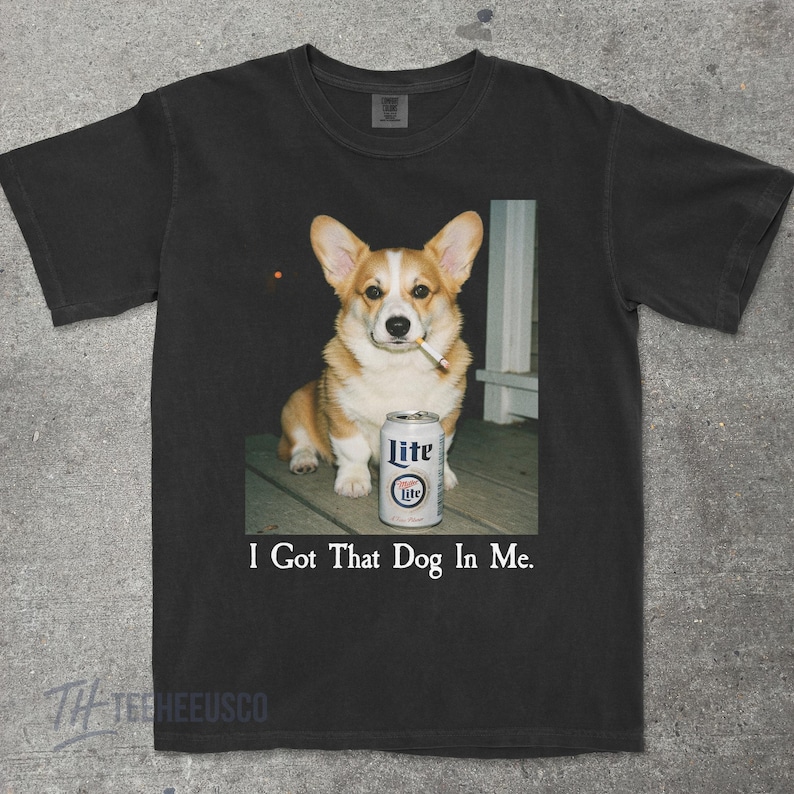 May include: Black t-shirt with a corgi dog smoking a cigarette, beside a can of Lite beer. The text "I Got That Dog In Me" is printed below the image. The shirt is made of a soft material. The image is printed on the front.
