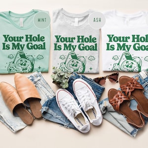 Może przedstawiać: Trzy koszulki w kolorze miętowym, popielatym i białym z napisem "Your Hole Is My Goal" i postacią kresk&oacute;wkowej piłki golfowej. Koszulki leżą na jasnobrązowej powierzchni z parą brązowych sandał&oacute;w, parą białych but&oacute;w sportowych i parą niebieskich dżins&oacute;w.