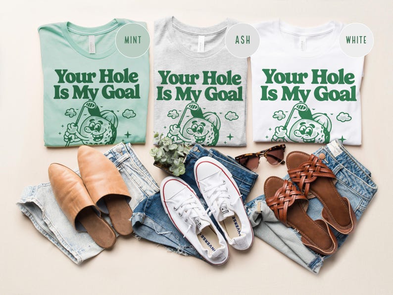 Może przedstawiać: Trzy koszulki w kolorze miętowym, popielatym i białym z napisem "Your Hole Is My Goal" i postacią kresk&oacute;wkowej piłki golfowej. Koszulki leżą na jasnobrązowej powierzchni z parą brązowych sandał&oacute;w, parą białych but&oacute;w sportowych i parą niebieskich dżins&oacute;w.