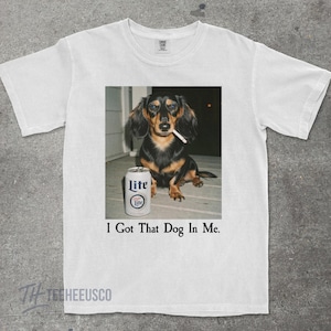 May include: White t-shirt featuring a graphic of a dachshund with a cigarette in its mouth and a can of Lite beer. The text "I Got That Dog In Me" is printed below the image.