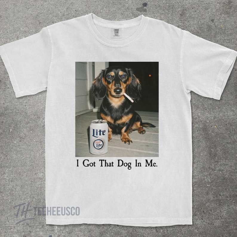 May include: White t-shirt featuring a graphic of a dachshund with a cigarette in its mouth and a can of Lite beer. The text "I Got That Dog In Me" is printed below the image.