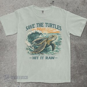 May include: Light blue t-shirt with a graphic of a sea turtle swimming in the ocean. The text "SAVE THE TURTLES" is above the turtle, and "- HIT IT RAW -" is below. The shirt has a crew neck and short sleeves.
