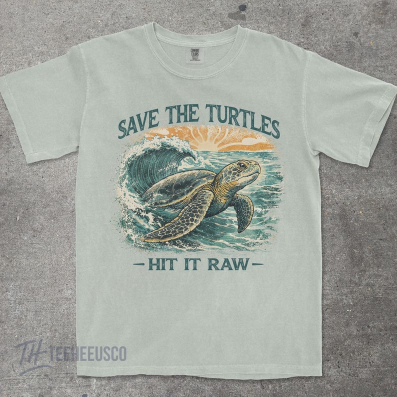 May include: Light blue t-shirt with a graphic of a sea turtle swimming in the ocean. The text "SAVE THE TURTLES" is above the turtle, and "- HIT IT RAW -" is below. The shirt has a crew neck and short sleeves.