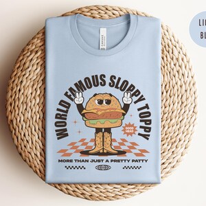 May include: Light blue t-shirt featuring a cartoon burger wearing cowboy boots and the text "World Famous Sloppy Toppy" and "More Than Just A Pretty Patty".
