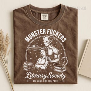May include: A brown t-shirt featuring a white graphic print of a horned creature standing behind a person with crossed arms. The design includes stacks of books, a flying saucer, stars, and the text "MONSTER FUCKERS Literary Society WE CAME FOR THE PLOT".