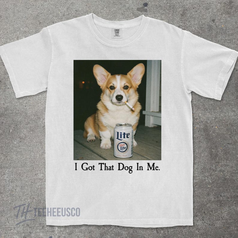 May include: White t-shirt with a square image of a corgi holding a cigarette, beside a Lite beer can. The text "I Got That Dog In Me" is printed below. The shirt has a crew neck and short sleeves. The image is printed on the front.