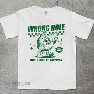 May include: White t-shirt with a green graphic of a golf ball character holding a golf club and giving a peace sign. The text "WRONG HOLE" and "BUT I LIKE IT ANYWAY" are above and below the graphic, respectively. The words "STILL COUNTS" are also included.