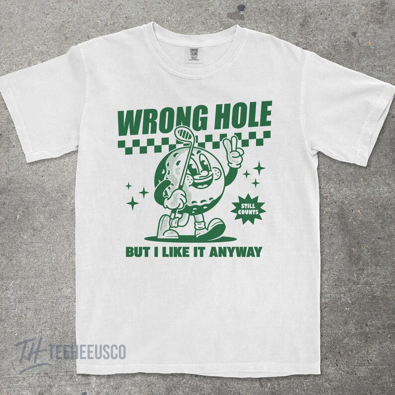 May include: White t-shirt with a green graphic of a golf ball character holding a golf club and giving a peace sign. The text "WRONG HOLE" and "BUT I LIKE IT ANYWAY" are above and below the graphic, respectively. The words "STILL COUNTS" are also included.
