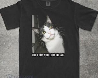 May include: Black t-shirt featuring a black and white cat with yellow eyes. The cat is looking at the viewer with the text "THE FUCK YOU LOOKING AT?" printed below the image. The brand logo is at the bottom.