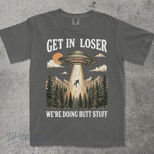 May include: Dark gray t-shirt with a graphic of a UFO beaming up a person over a forest and mountains. The text reads "GET IN LOSER" above the UFO and "WE'RE DOING BUTT STUFF" below.