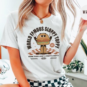 May include: White t-shirt featuring a cartoon burger graphic with the text "World Famous Sloppy Toppy" and "More Than Just A Pretty Patty".